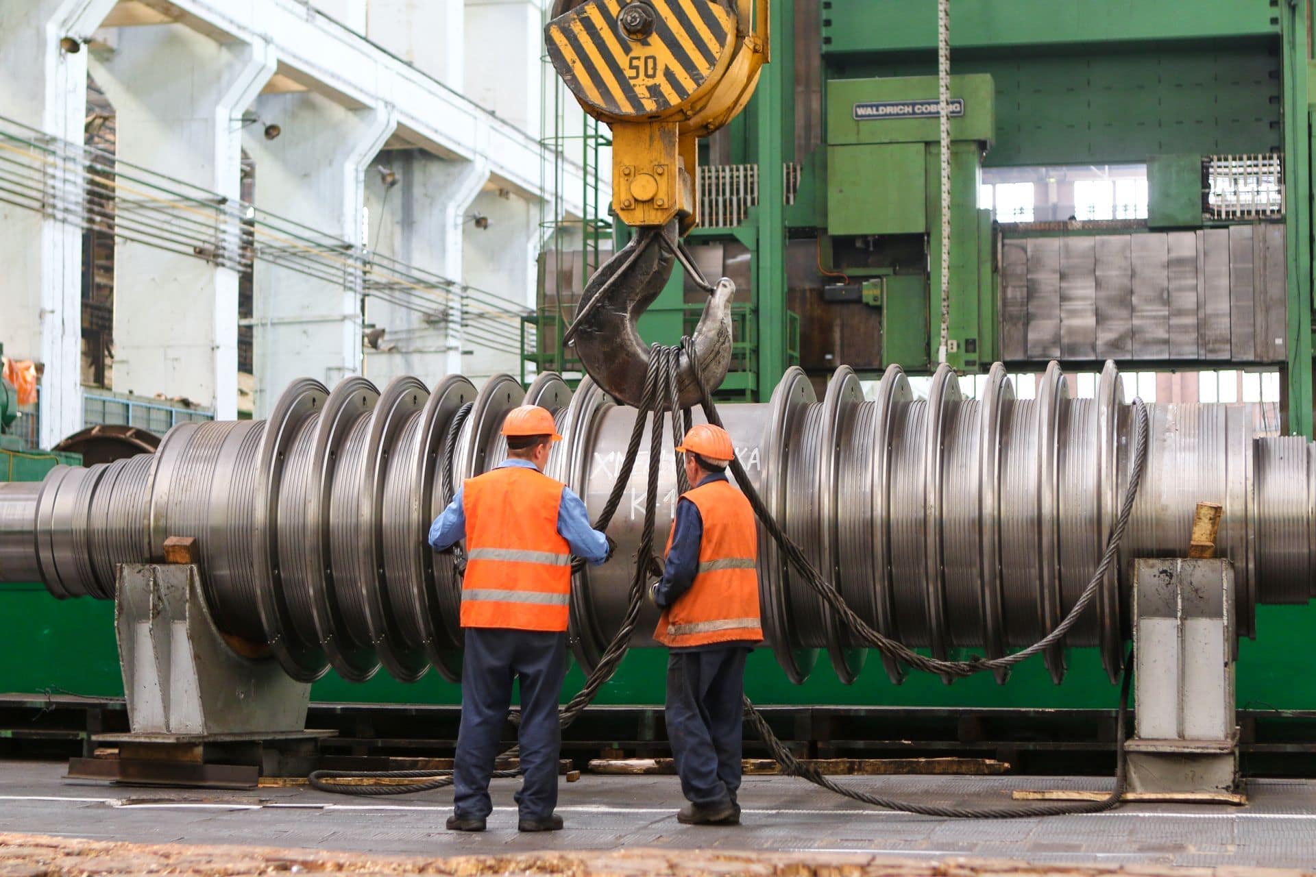 Due operai guidano un grande rotore di turbina appeso a una gru in un capannone di costruzione macchine