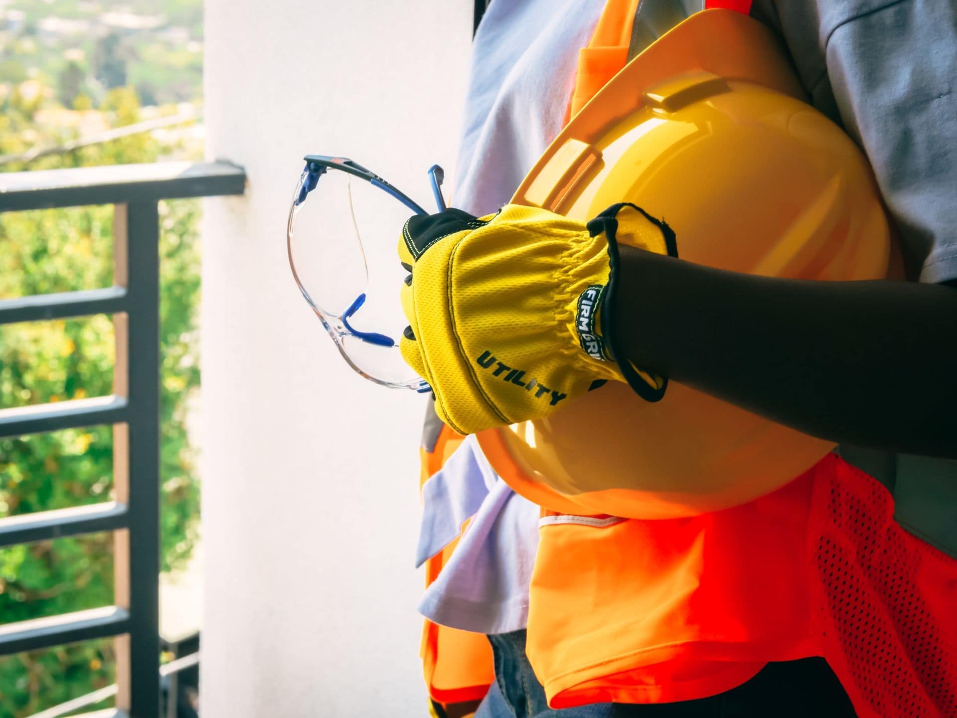 Worker in a high-visibility vest holding a hard hat, safety glasses and protective gloves